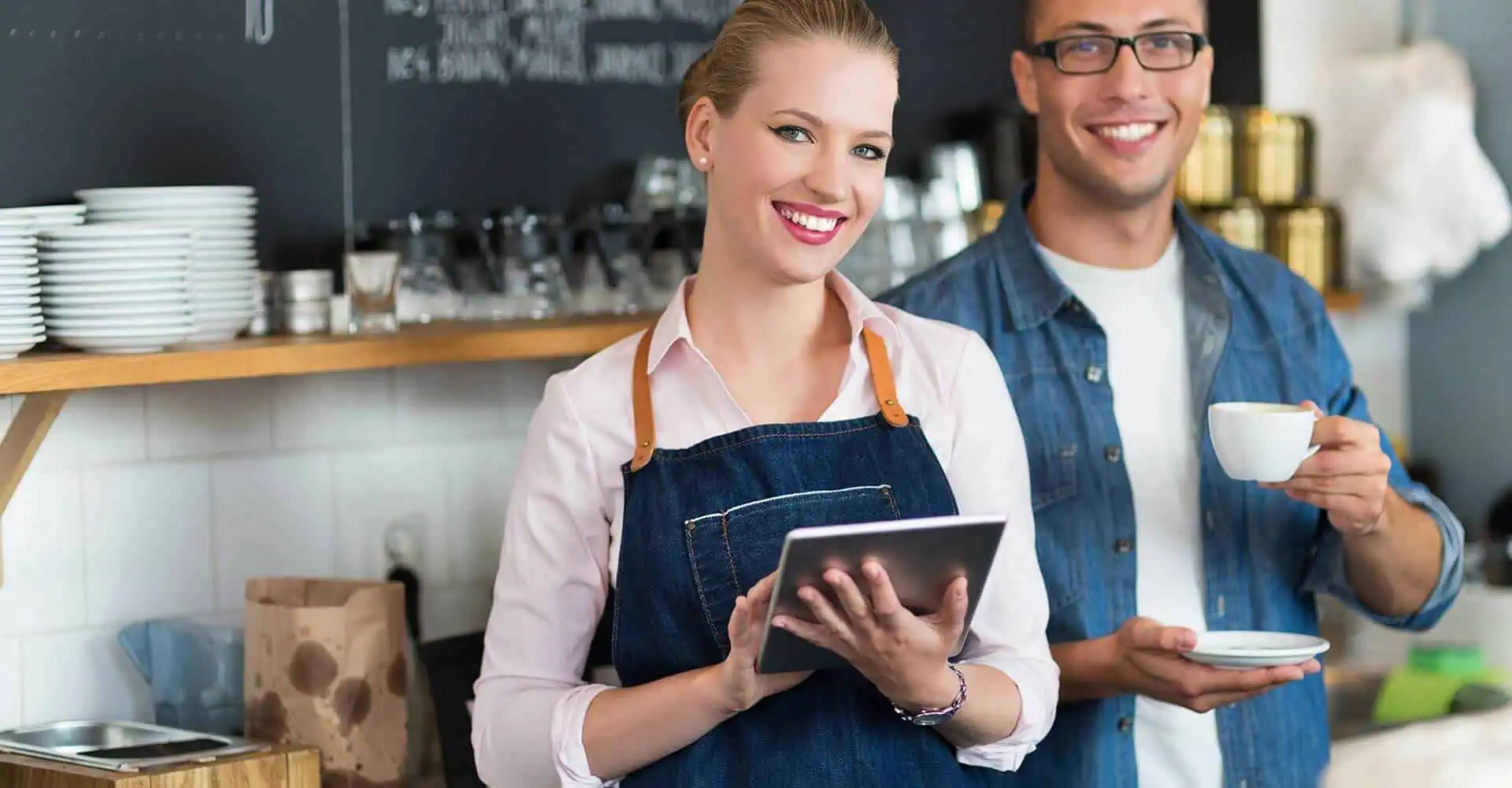 Two people in a cafe use a tablet POS system for seamless credit card payment processing, highlighting merchant services and payment solutions with shelves behind them.