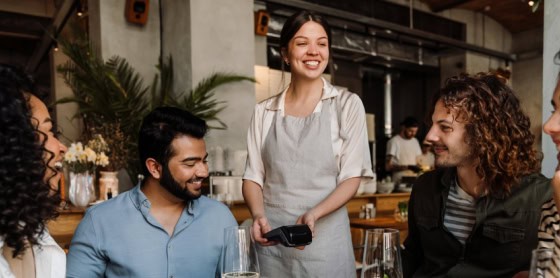 In a lively restaurant, a server with a POS system offers payment solutions to diners.