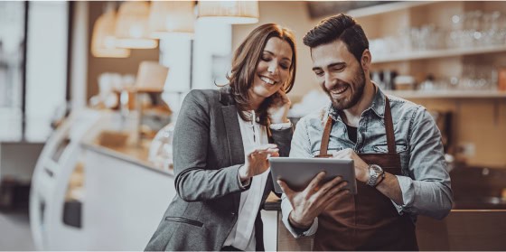 A woman and man in aprons smile, discussing POS and payment solutions on tablet.